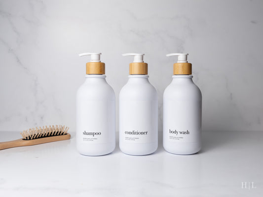 Set of 3 white shampoo, conditioner and body wash containers with bamboo pump on a bathroom counter with a hair brush