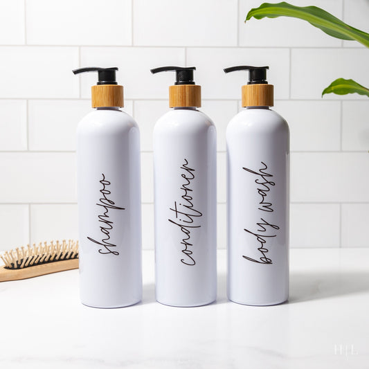 set of 3 white pump bottle dispensers with cursive script labels for shampoo, conditioner and body wash with black bamboo pump on a bathroom counter next to a hair brush and plant
